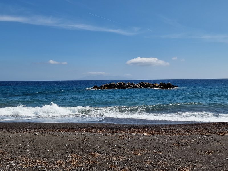 A quiet stretch of black volcanic sand beach near Kamari with a small rocky outcrop in the sea
