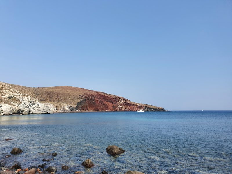 Red Beach's rust-colored cliffs and turquoise water, with a sailing boat anchored in the bay