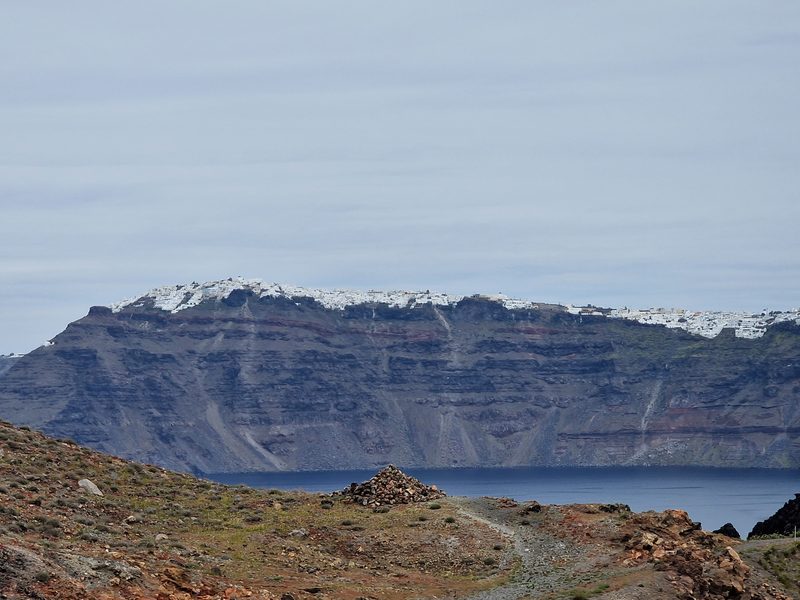 A dusty path marked by a stone cairn crosses the bare volcanic ground of Nea Kameni, with the caldera and Santorini's clifftop town in the background