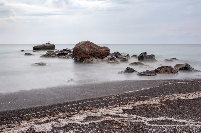 A serene, black sand beach with dramatic cliffs at Vlychada Beach in Exomitis, Santorini, Greece.